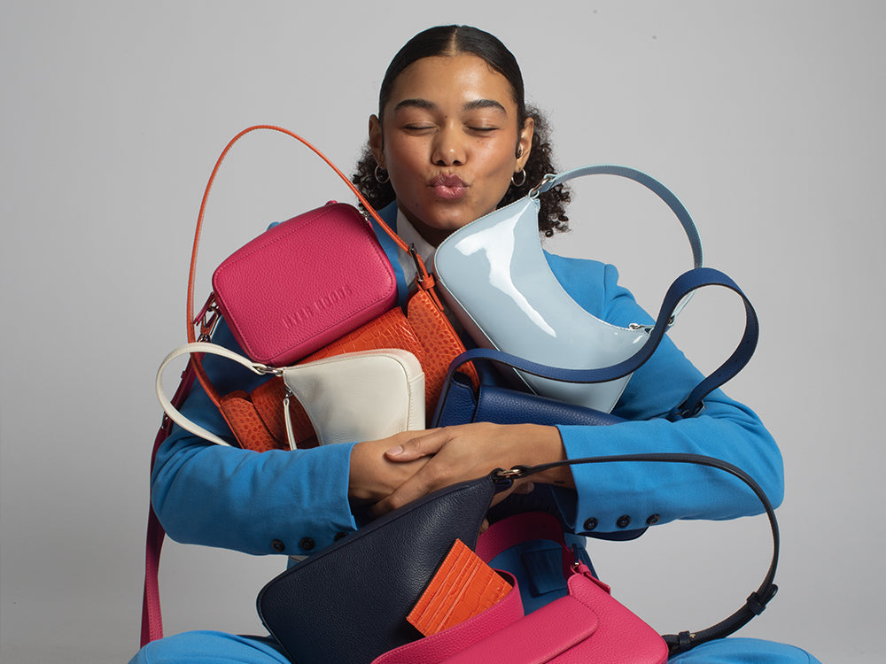a girl in a bright blue suit sits cross-legged, her arms clutches around a large pile of colorful handbags like a hug