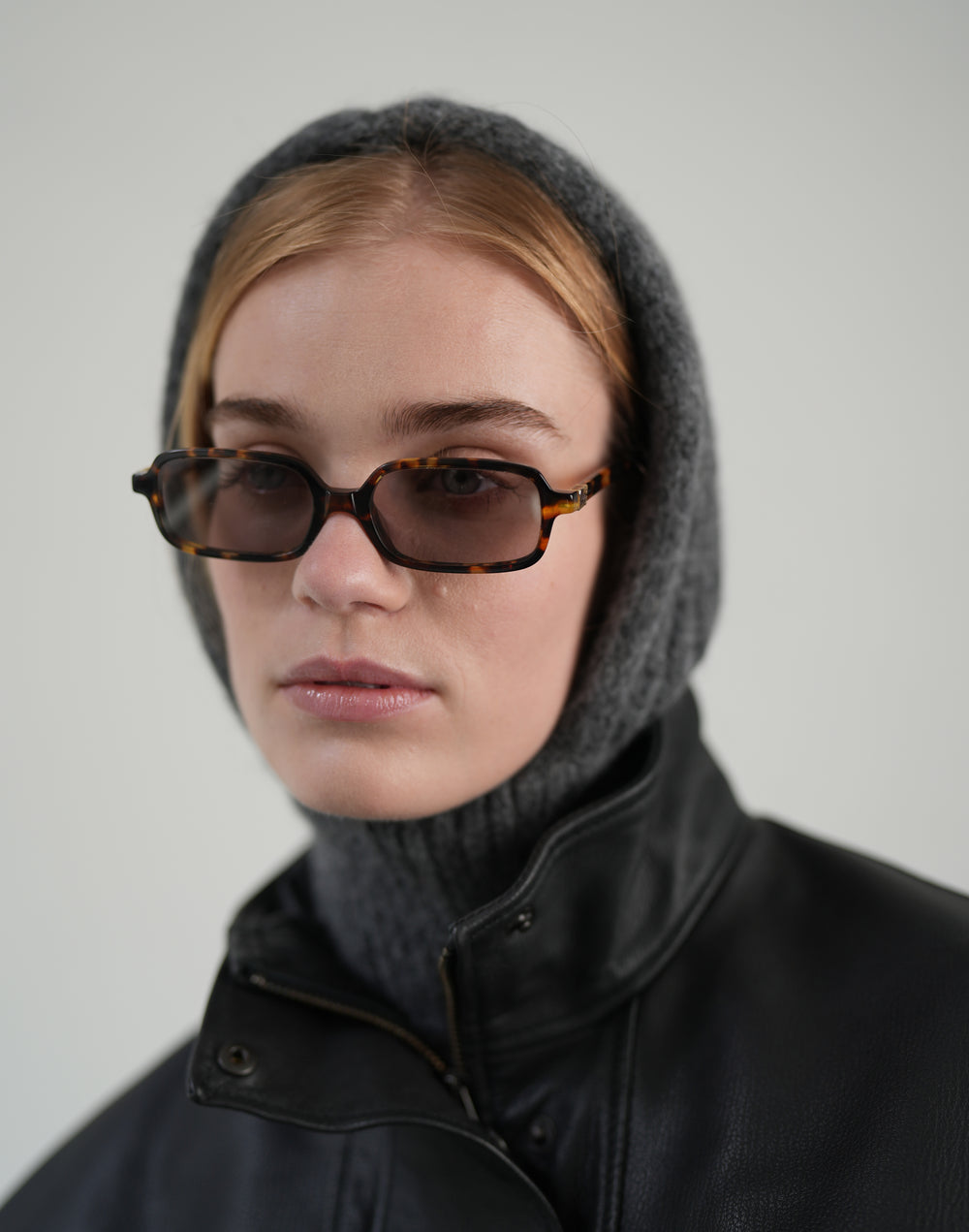 A model wearing rectangular tortoiseshell sunglasses with dark tinted lenses, styled with a gray knit hood and a black leather jacket against a plain light gray background. #color_grey