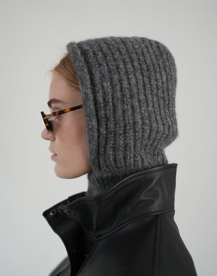 A side-profile view of a person wearing a dark gray ribbed knit hooded balaclava with a high neck, styled with tortoiseshell glasses and a black leather jacket against a light gray background. #color_grey