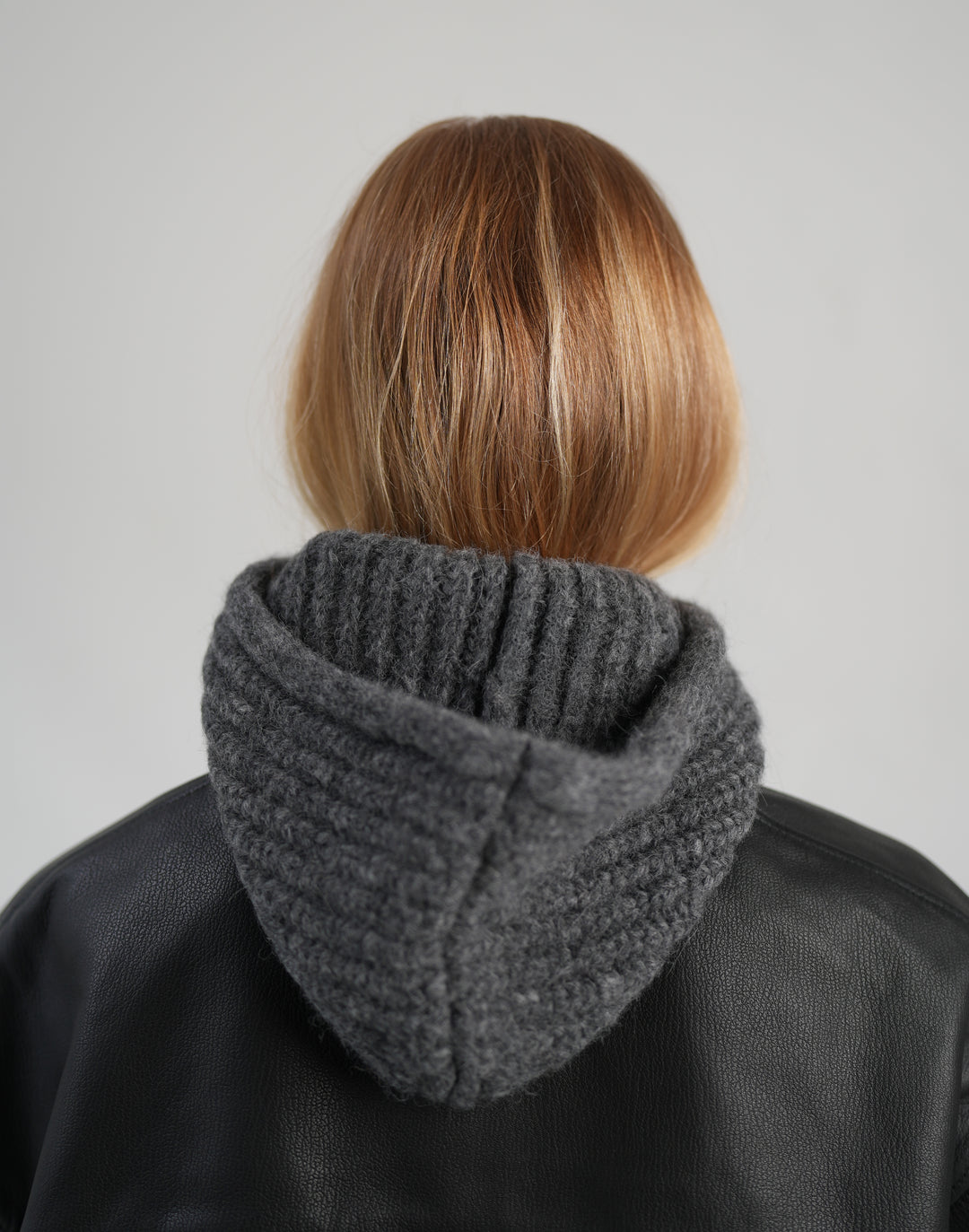 Back view of a person with short blonde hair wearing a dark gray ribbed knit hooded balaclava under a black leather jacket, against a light gray studio background. #color_grey