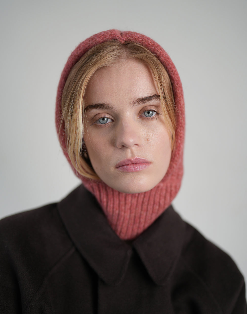 A close-up portrait of a model wearing a dusty rose pink ribbed knit hooded neck warmer over a dark brown coat, set against a plain light gray background. #color_dusty-pink