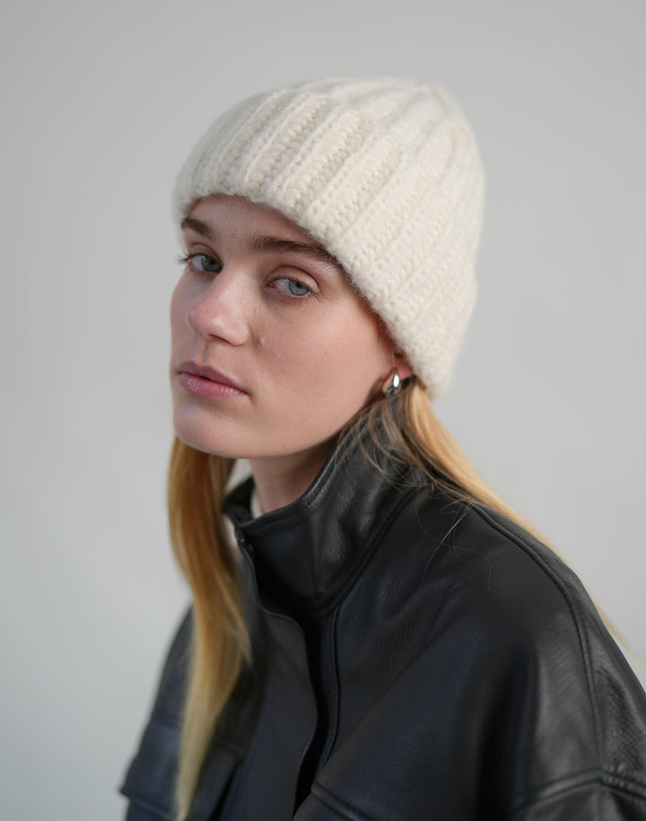 a cropped photo of a woman wearing a black leather funnel neck coat and a cream chunky wool ribbed beanie #color_cream