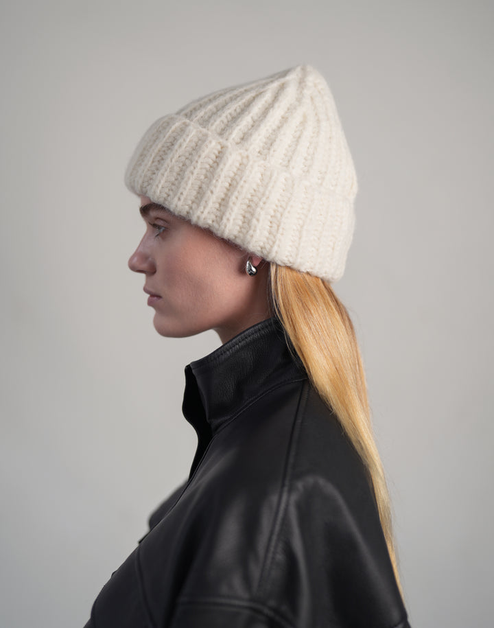 A woman in profile wearing a cream-colored ribbed knit beanie and a black leather jacket, photographed against a neutral gray studio background. #color_cream