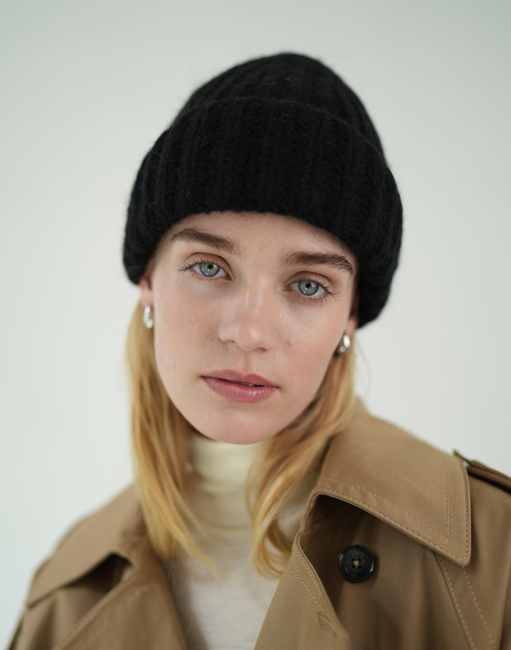 A woman wearing a black chunky ribbed knit beanie, a tan trench coat, and a cream turtleneck against a light gray studio background. #color_black