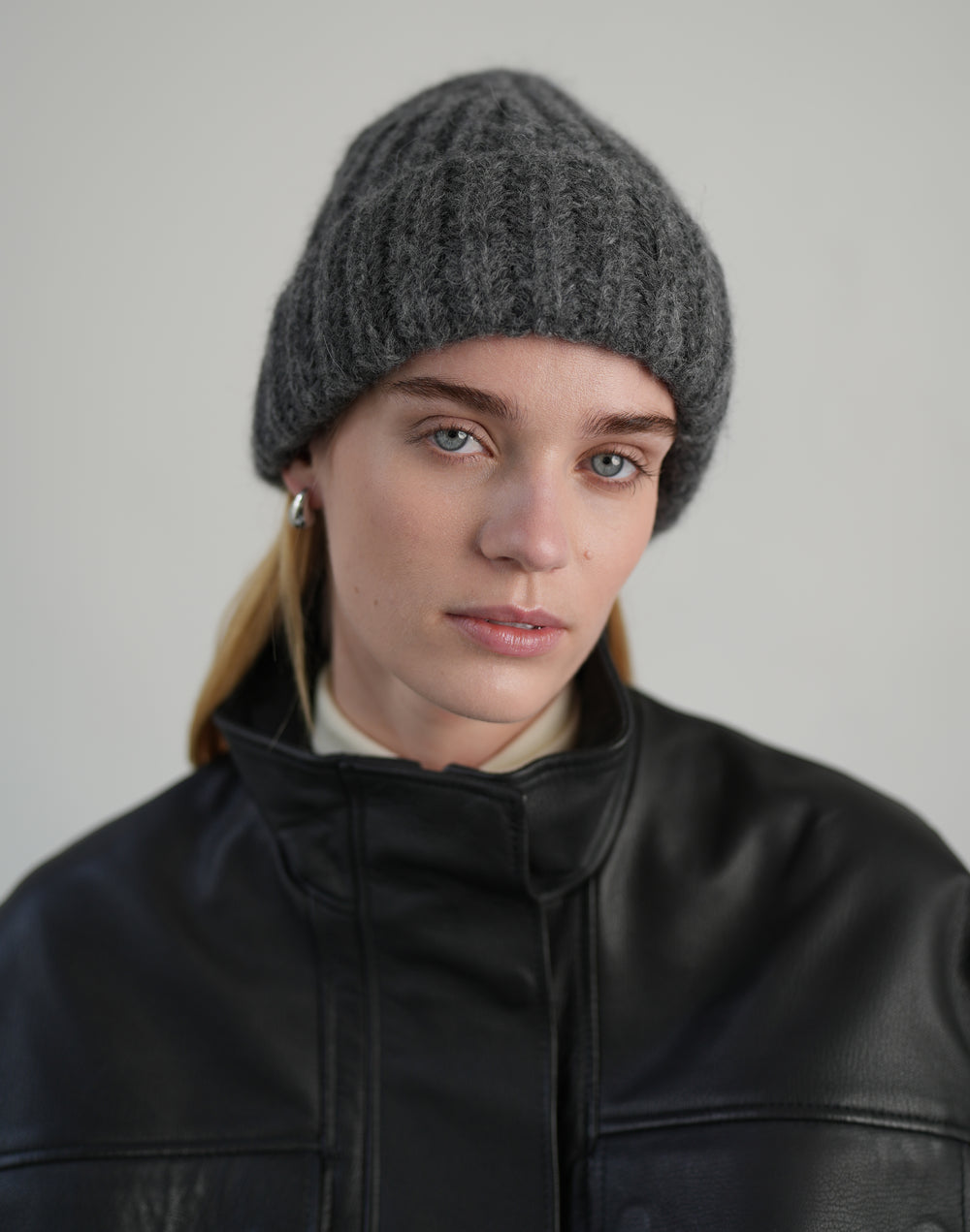 A woman wearing a dark gray ribbed knit beanie and a black leather jacket, photographed against a neutral light gray background. #color_grey