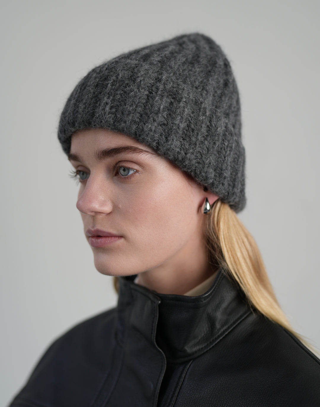 A woman in profile wearing a dark gray ribbed knit beanie and a black leather jacket, photographed against a neutral light gray background. #color_grey