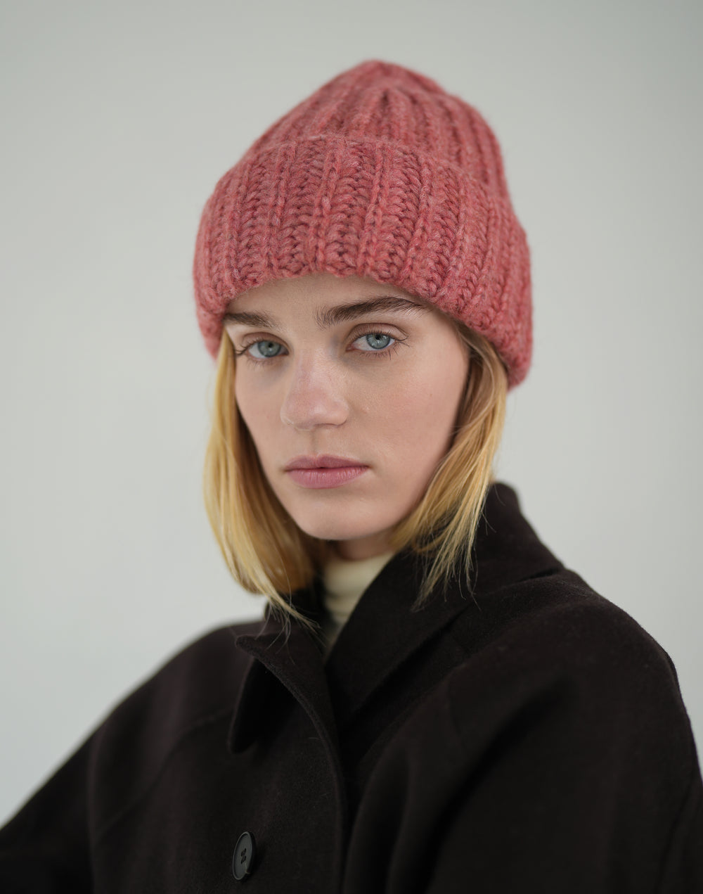 A woman wearing a chunky knit beanie in dusty rose pink and a dark brown coat, photographed against a neutral light gray studio background. #color_dusty-pink