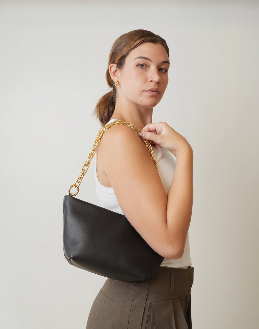 A woman wearing a white tank top and brown pants carries a black leather shoulder bag with a chunky gold chain strap against a plain light gray background.#color_gold