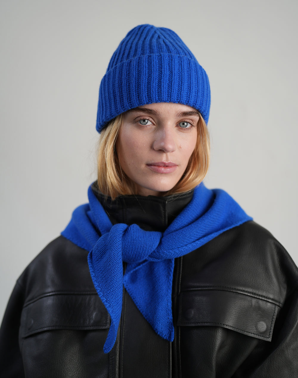 A woman wearing a bright royal blue ribbed knit beanie and a matching blue scarf, paired with a black leather jacket, against a neutral light gray background. #color_cobalt