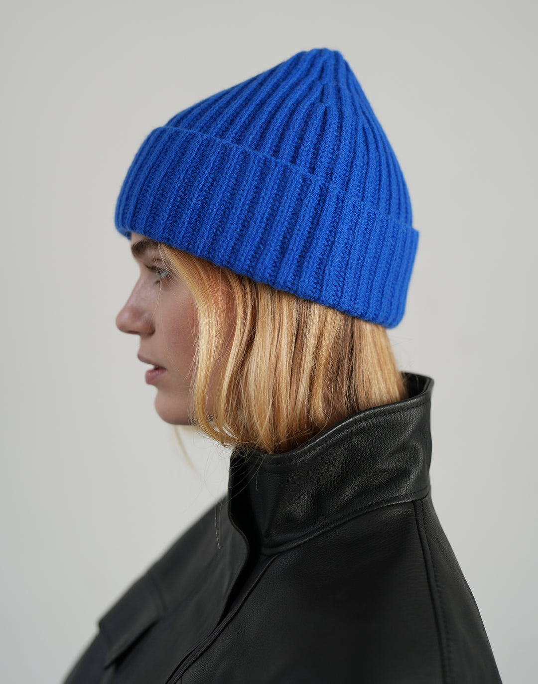 A woman in profile wearing a bright royal blue ribbed knit beanie and a black leather jacket, photographed against a plain light gray studio background. #color_cobalt