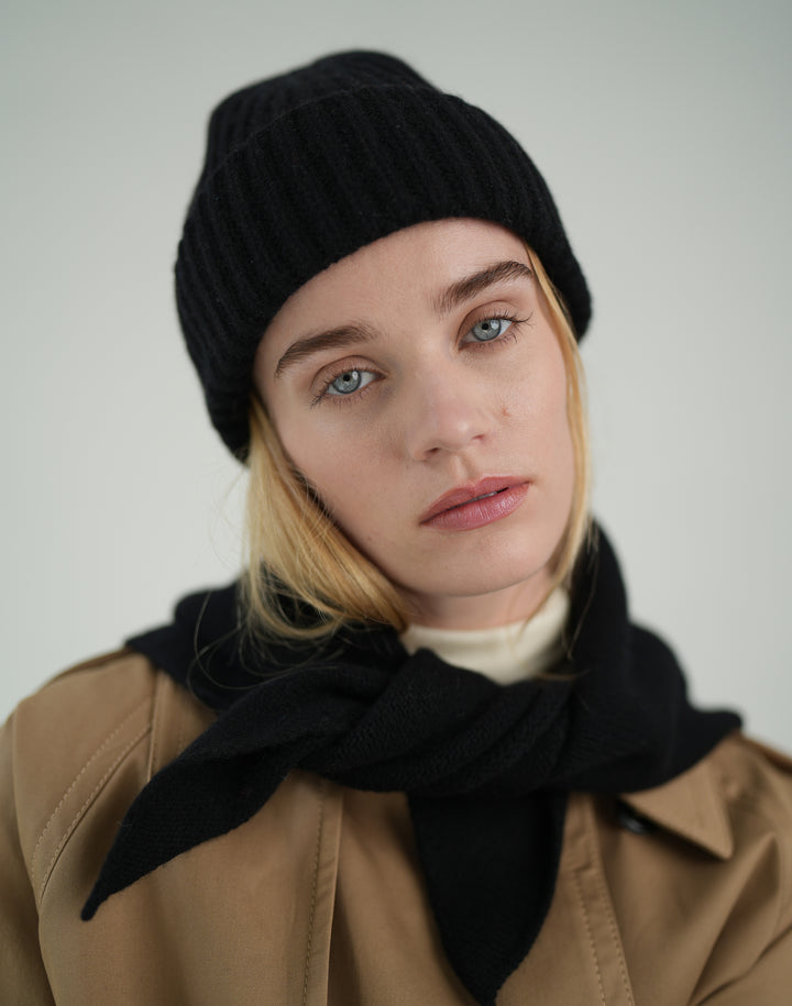 a close up image of a woman with blond hair wearing a black ribbed wool beanie, a black neck scarf, a white turtleneck and a khaki trench #color_black