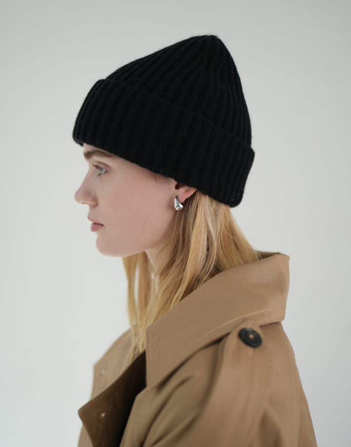 A woman in profile wearing a black ribbed knit beanie and a tan trench coat, photographed against a plain light gray studio background.#color_black