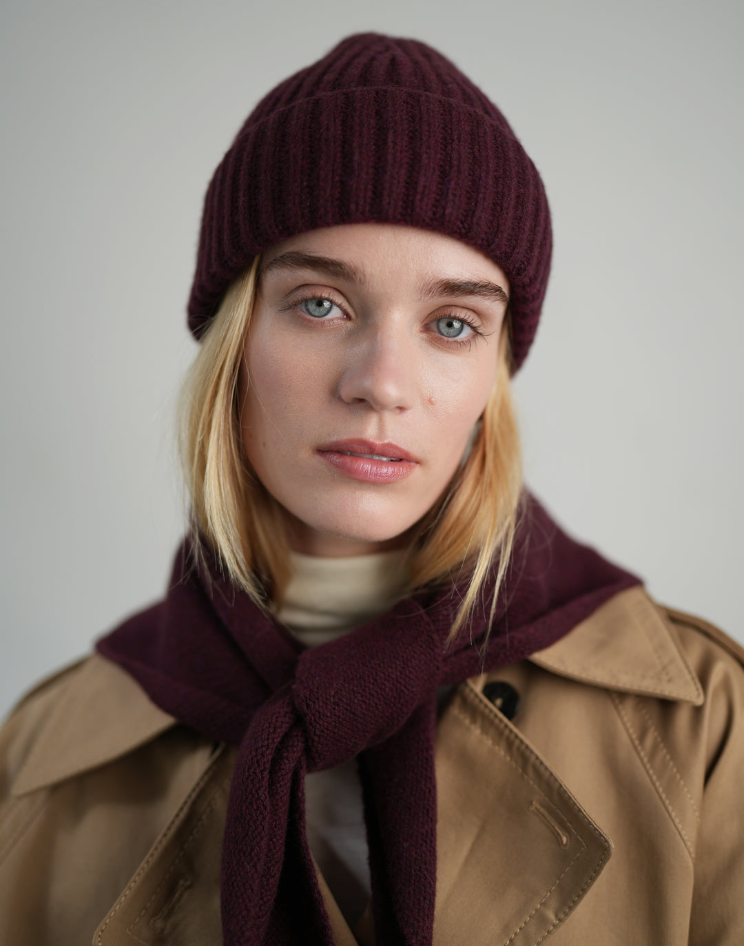 A woman wearing a deep burgundy ribbed knit beanie, a matching burgundy knit scarf, and a tan trench coat, photographed against a neutral light gray background.#color_bordeaux