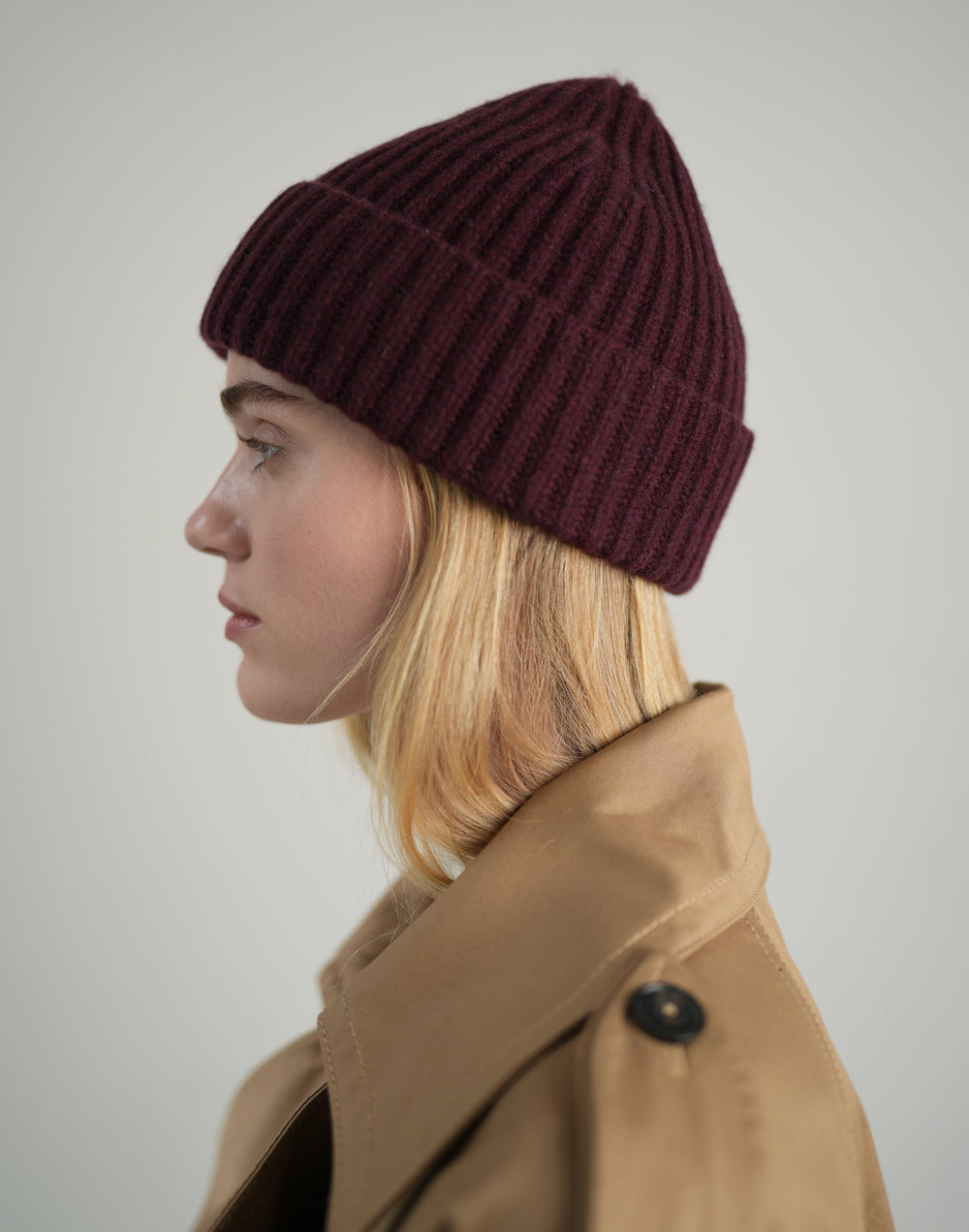 A cropped profile of a woman wearing a bordeaux knit ribbed beanie and a trench coat against a grey background #color_bordeaux