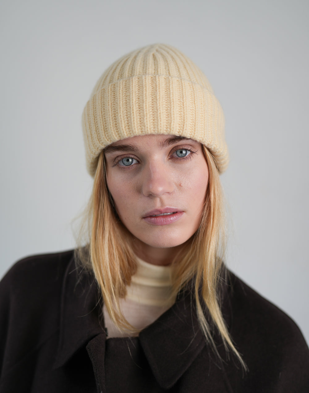A woman with blonde hair wearing a light yellow-colored ribbed knit beanie and a dark brown coat, photographed against a neutral light gray background.#color_butter-yellow