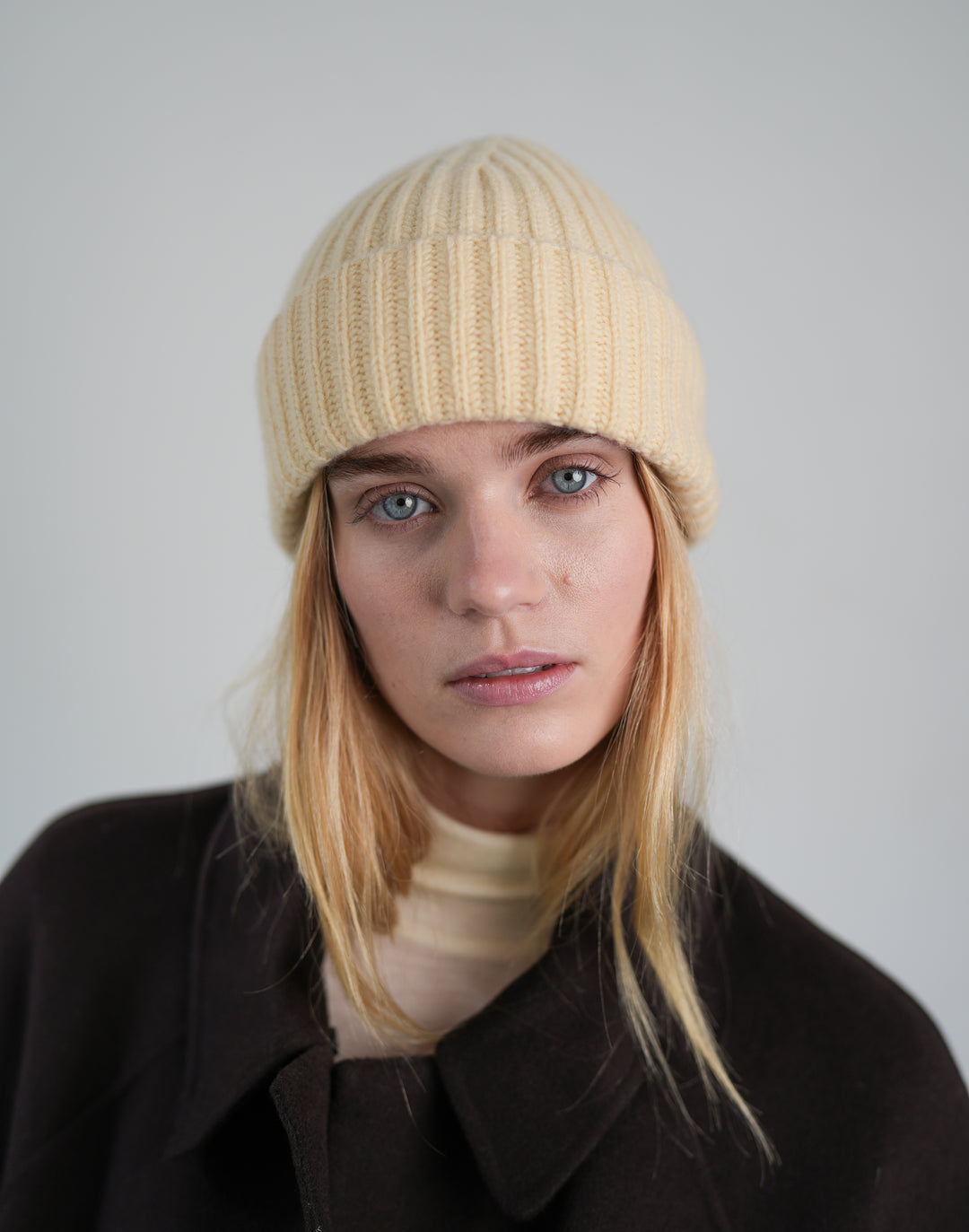 A woman with blonde hair wearing a light yellow-colored ribbed knit beanie and a dark brown coat, photographed against a neutral light gray background.#color_butter-yellow