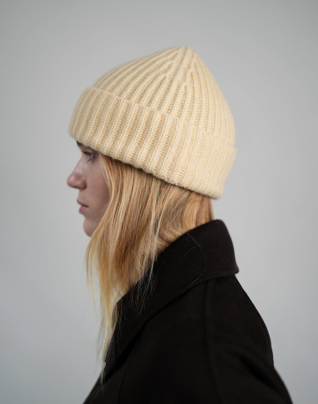 A woman in profile wearing a light yellow-colored ribbed knit beanie and a dark coat, photographed against a neutral light gray background. #color_butter-yellow
