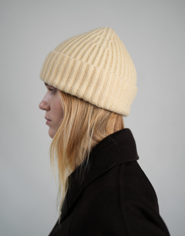 A woman in profile wearing a light yellow-colored ribbed knit beanie and a dark coat, photographed against a neutral light gray background. #color_butter-yellow