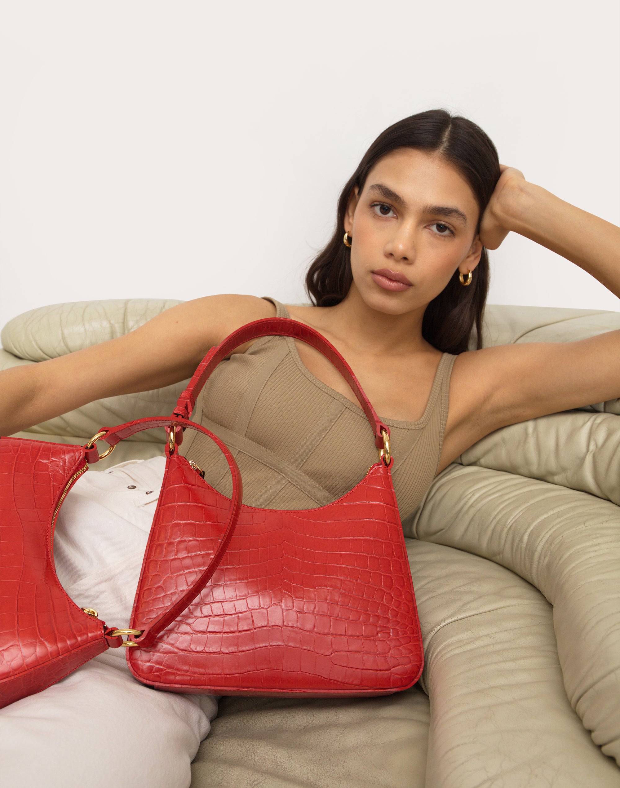 a woman lays on a khaki quilted couch in a khaki top holding a bright red croc medium shoulder bag 