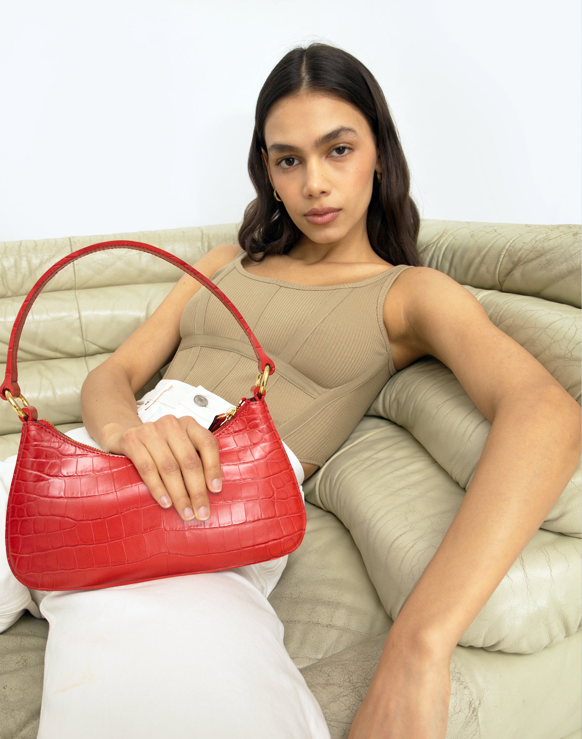 a woman lounging on a beige vintage chair wears a tan top, white pants and a bright red croc mini shoulder bag.