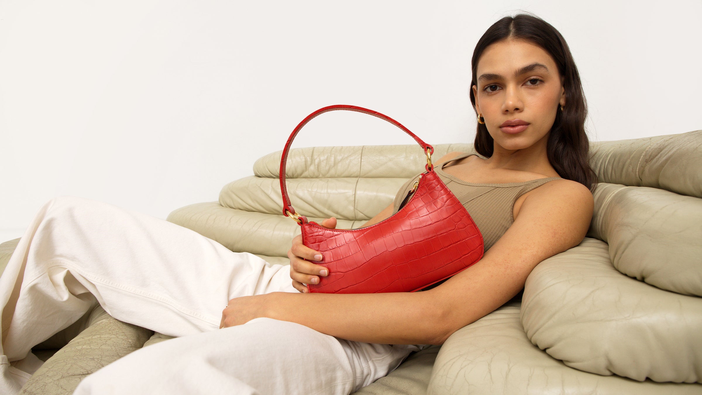 a woman lays on a khaki quilted couch in a khaki top holding a bright red croc mini shoulder bag