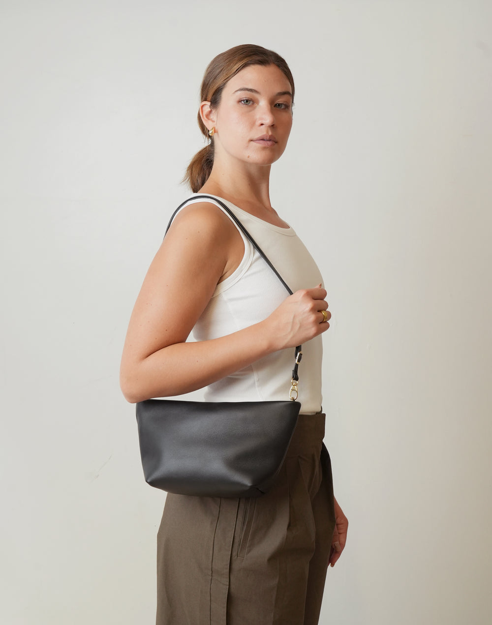 A woman wearing a white tank top and brown trousers carries a HYER GOODS  black leather La Sala shoulder bag with a thin black strap and gold-tone hardware against a plain light gray background. #color_black-gold