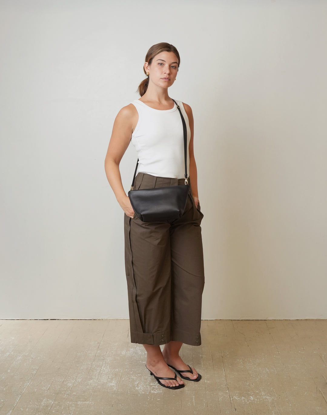 A woman stands in a studio wearing a white sleeveless top, wide-leg brown cropped pants, and black flip-flops, while carrying a HYER GOODS La Sala minimal black leather crossbody bag #color_black-gold