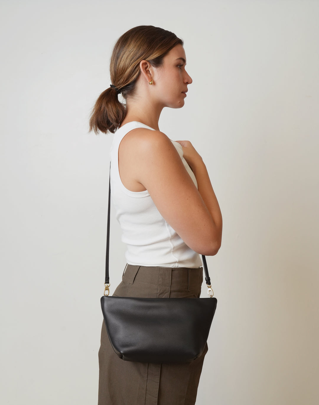 A woman in a white tank top and brown pants is shown in profile wearing a black leather crossbody shoulder bag with a thin black strap and gold-tone hardware against a plain light gray background. #color_black-gold