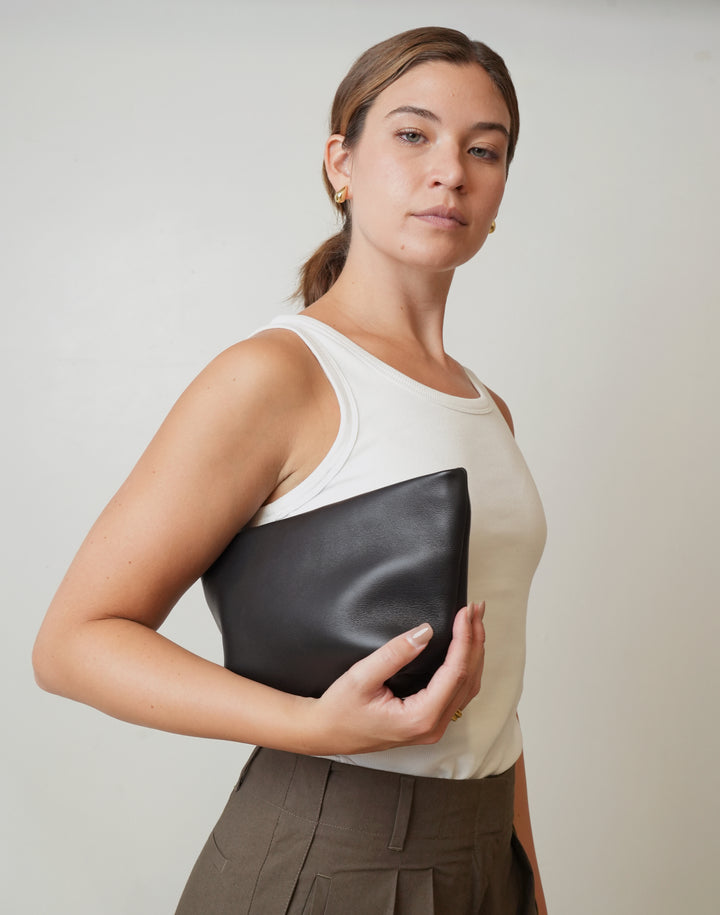 A woman wearing a white ribbed tank top and brown pleated trousers holds a black leather clutch bag against her torso, posed against a neutral light gray background. #color_black-gold