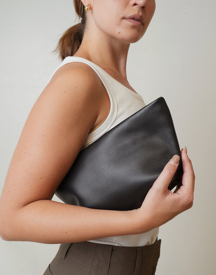 A woman in a white tank top holds a large, soft black leather clutch bag against her torso, shown from the side against a plain light gray background. #color_black-gold
