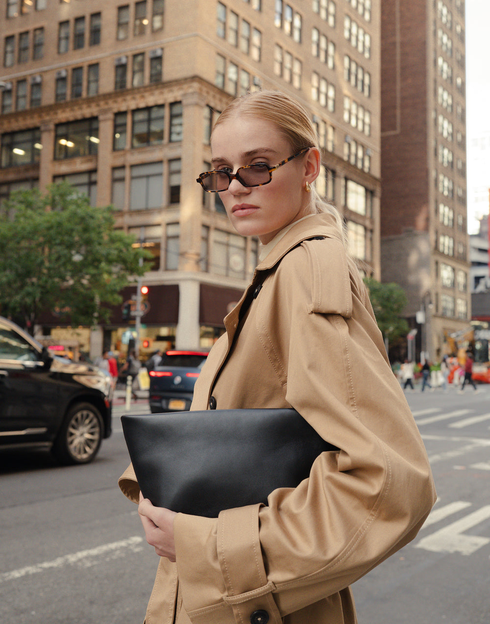 a cropped photo of a woman standing in the streets of NYC, sunglasses hanging on her nose, in a trench coat, clutching a black leather La Sala Clutch under her arm while cars drive behind her #color_black-silver