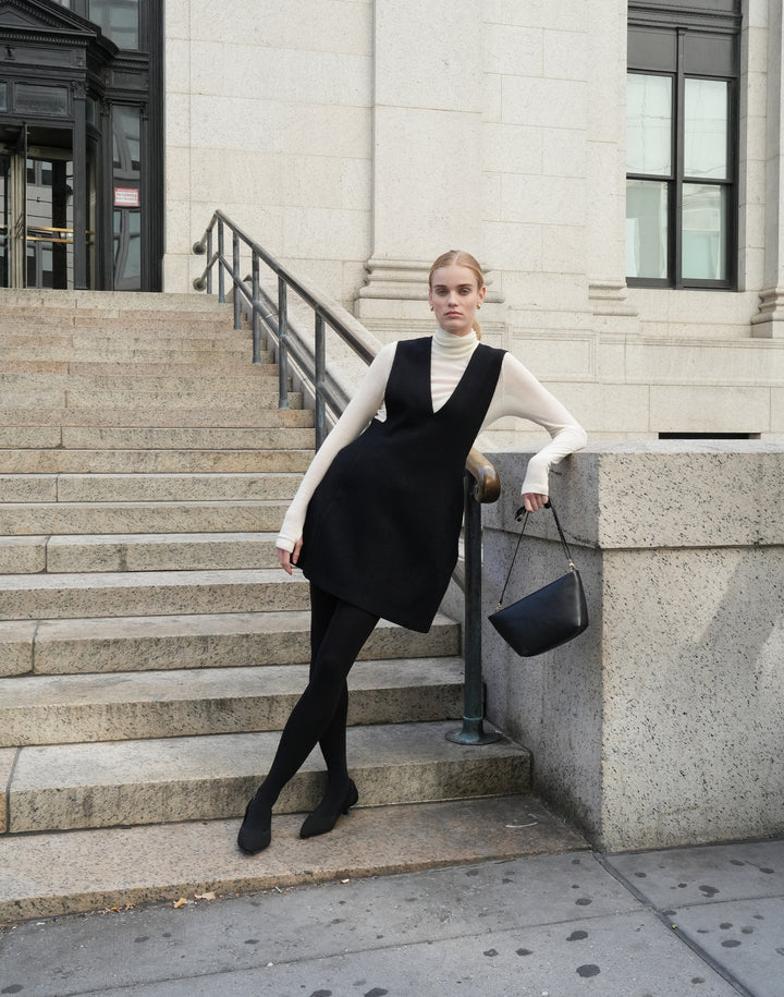 a woman in a sheer cream turtleneck, black v neck mini dress, tights and pointy kitten heels leans agains a nyc building holding a black shoulder bag with a thing strep from her fingertips.  #color_black-gold