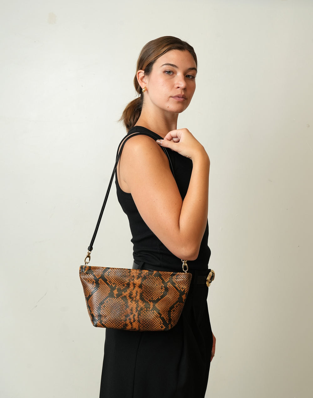 A woman wearing a black sleeveless dress carries a HYER GOODS La Sala brown and black snakeskin-patterned shoulder bag with a black strap against a plain off-white background.  #color_tan-python