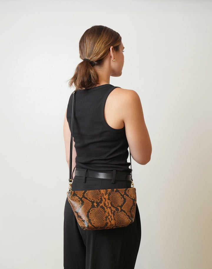 A woman shown from the back wearing a black sleeveless top and black pants, carrying a HYER GOODS La Sala brown and black snakeskin-patterned crossbody bag against a plain light gray studio background.  #color_tan-python