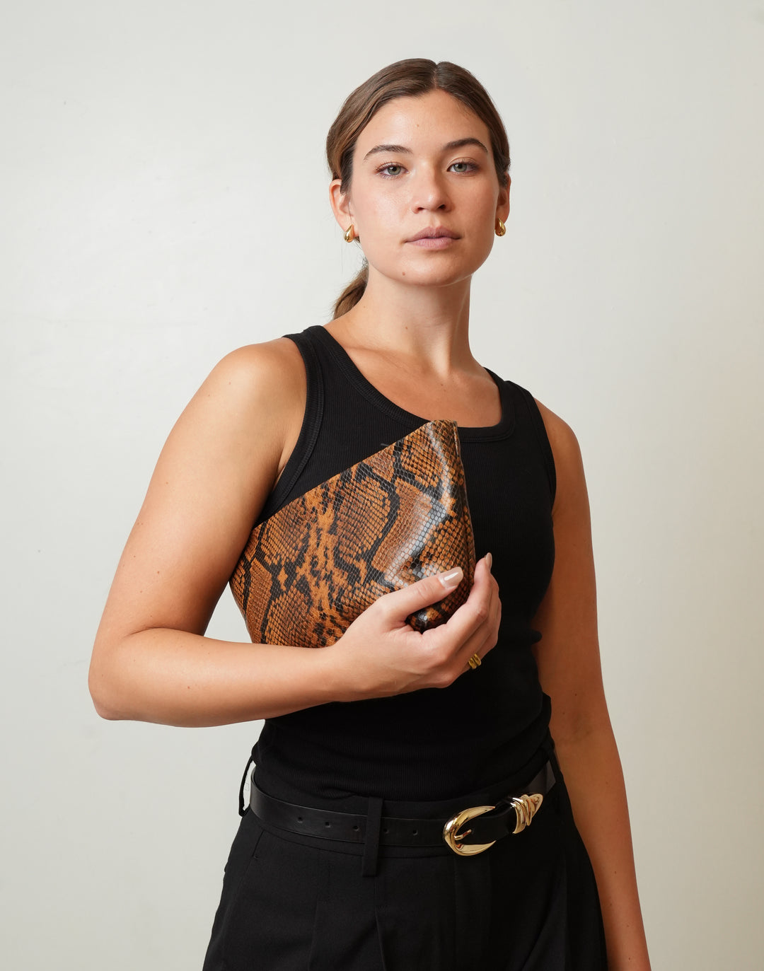 A woman wearing a black sleeveless top and black trousers holds a black clutch bag with a  HYER GOODS La Sala brown and black snakeskin pattern against a light gray background.  #color_tan-python