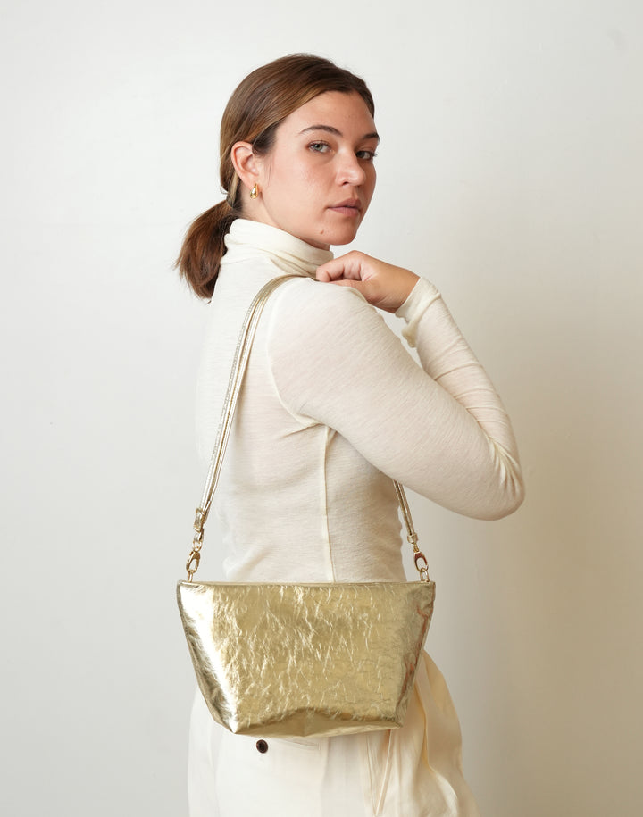 A woman wearing a cream-colored turtleneck sweater and cream skirt, carrying a HYER GOODS LA SALA metallic gold crinkled leather shoulder bag against a plain off-white background.#color_gold