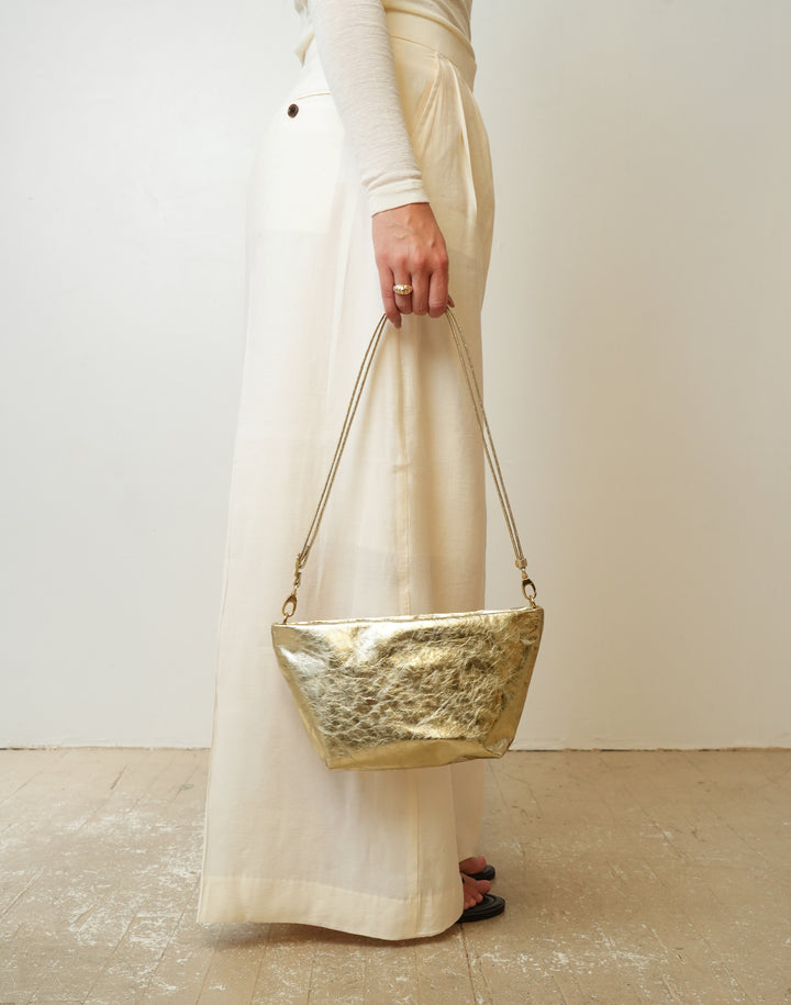 A person wearing a cream long-sleeve top and matching wide-leg skirt holds a  HYER GOODS LA SALA textured metallic gold shoulder bag with a thin gold chain strap against a plain off-white wall and light wood floor.#color_gold