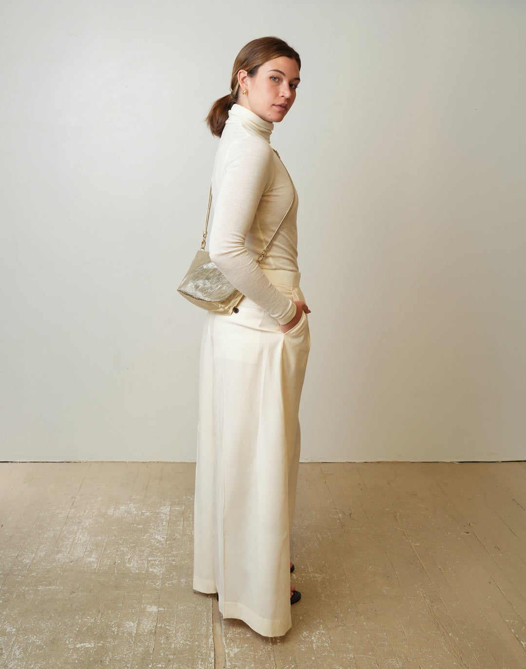 A woman stands in profile wearing a cream turtleneck top and matching wide-leg skirt, holding a small HYER GOODS LA SALA metallic crossbody bag against a plain off-white wall and light wood floor.#color_gold