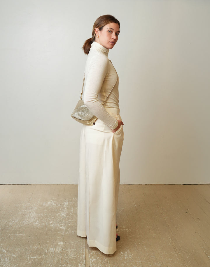 A woman stands in profile wearing a cream turtleneck top and matching wide-leg skirt, holding a small HYER GOODS LA SALA metallic crossbody bag against a plain off-white wall and light wood floor.#color_gold
