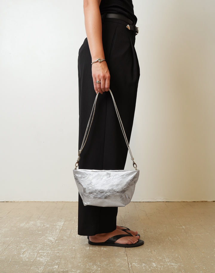 A person wearing black wide-leg trousers and black sandals holds a HYER GOODS La Sala crinkled metallic silver shoulder bag with a thin silver chain strap against a plain beige wall and light floor.  #color_silver