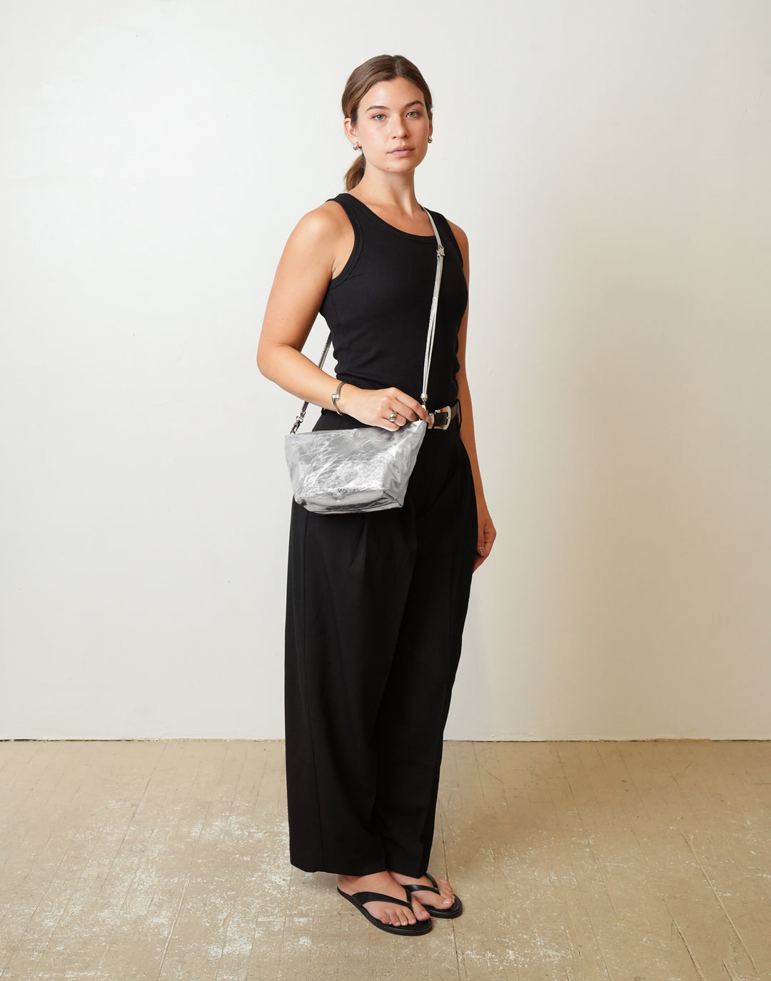 A woman modeling a black sleeveless jumpsuit with wide-leg pants, carrying a HYER GOODS La Sala shiny silver crossbody bag, standing against a plain off-white wall. #color_silver