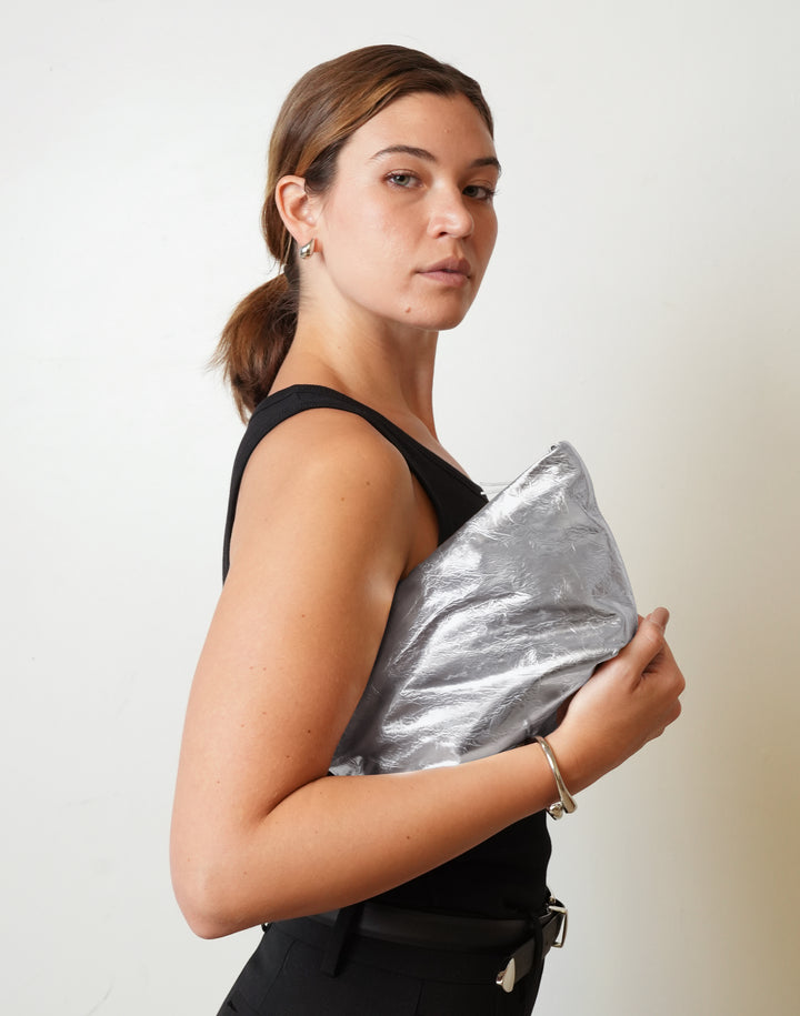 A woman with her hair in a ponytail, wearing a black sleeveless top and black pants, holds a HYER GOODS La Sala shiny silver metallic clutch bag against her side, photographed against a plain off-white background. #color_silver