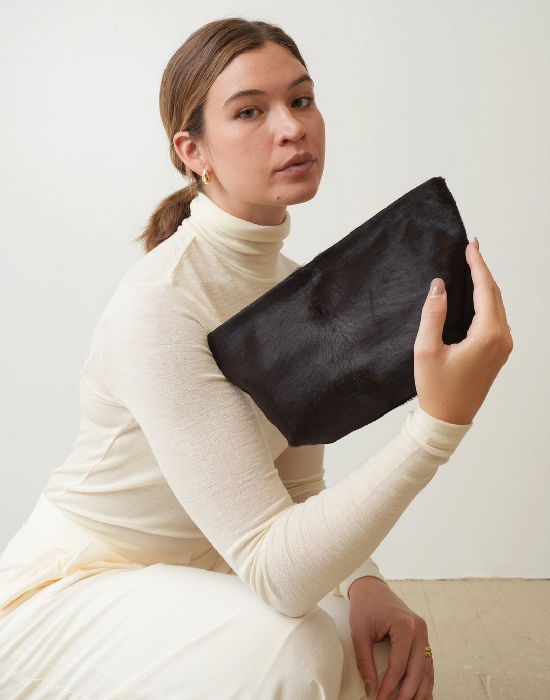 A woman in a cream ribbed turtleneck sweater and matching pants holds a brown pony hair clutch bag against a plain off-white background.HYER GOODS La Sala mini convertible shoulder bag #color_dark-chocolate-pony