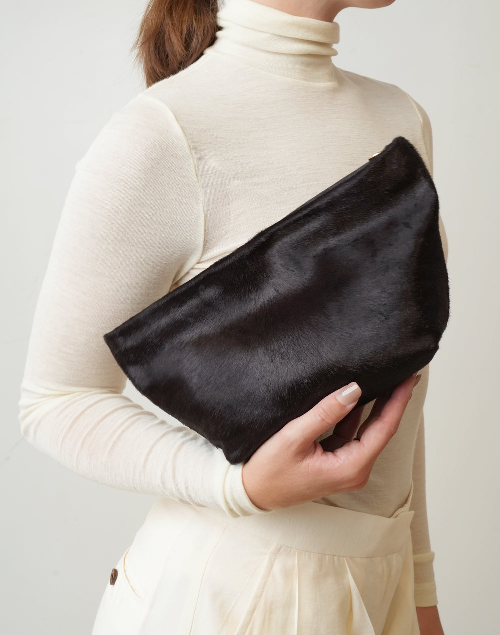 A person wearing a cream turtleneck sweater holds a brown pony hair clutch bag against a plain light background. HYER GOODS La Sala mini convertible clutch #color_dark-chocolate-pony