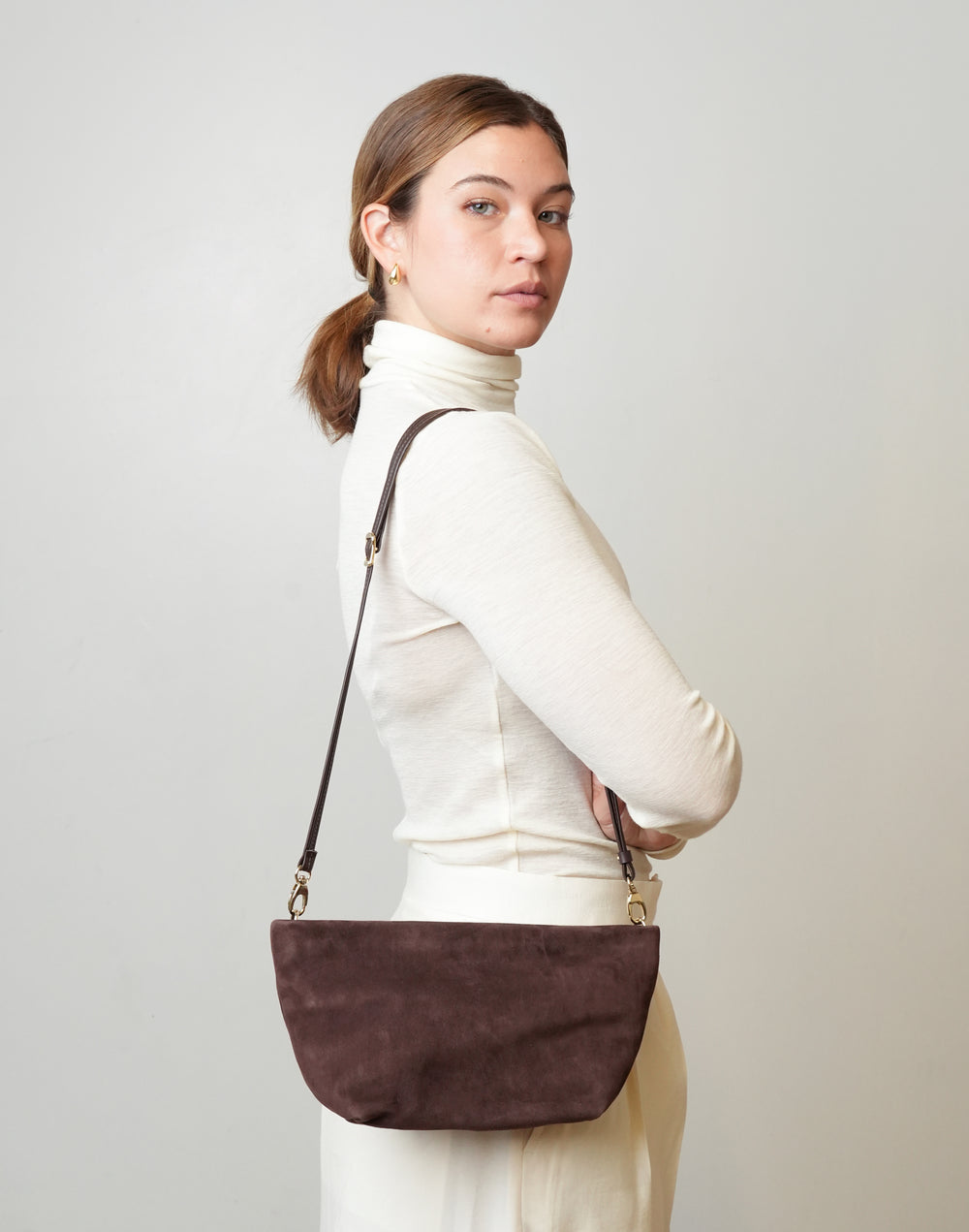 A woman wearing a cream turtleneck sweater and cream skirt, holding a HYER GOODS La Sala dark brown suede half-moon shoulder bag with a thin dark strap and gold-tone hardware, against a light gray studio background #color_chocolate-suede
