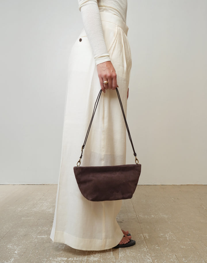 A person wearing a cream long-sleeve top and matching wide-leg skirt holds a HYER GOODS La Sala dark brown suede shoulder bag with a thin black strap and gold-tone hardware against a plain off-white wall and concrete floor. #color_chocolate-suede