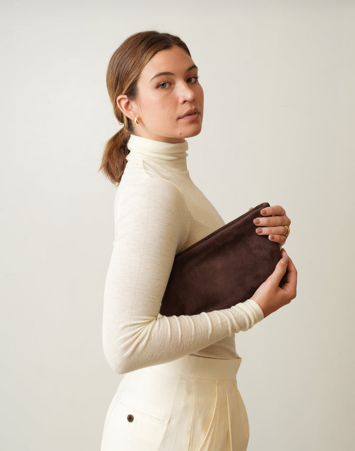 A woman in a cream turtleneck sweater and matching pants holds a HYER GOODS la sala dark brown suede clutch bag against her side, photographed against a plain light beige background. #color_chocolate-suede