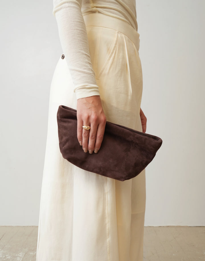 A person wearing a cream long-sleeve top and matching wide-leg skirt holds a HYER GOODS la sala dark brown  suede clutch bag against a plain off-white wall. #color_chocolate-suede