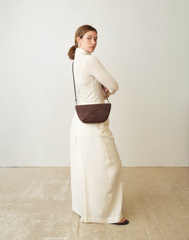 A woman in a cream-colored long-sleeve maxi dress stands in profile, wearing a HYER GOODS la sala dark brown crescent-shaped crossbody bag with a thin black strap, set against a minimalist off-white wall and light wooden floor. #color_chocolate-suede