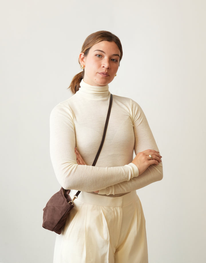 A woman wearing a cream ribbed turtleneck sweater and matching cream trousers, carrying a HYER GOODS la sala small brown crossbody bag, posed against a light off-white studio background. #color_chocolate-suede